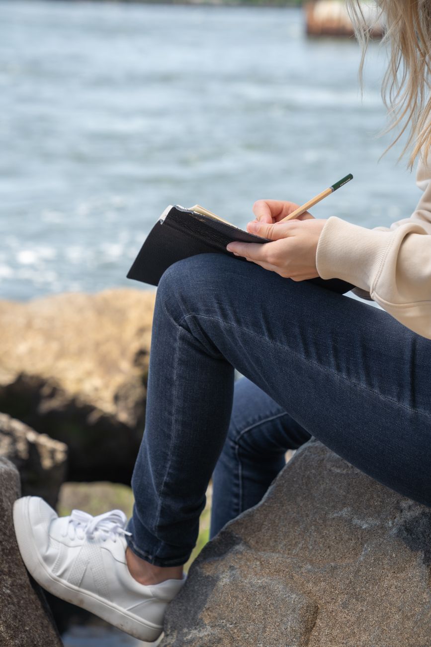 A woman sitting on rocks by the water, writing in a notebook with a pencil.